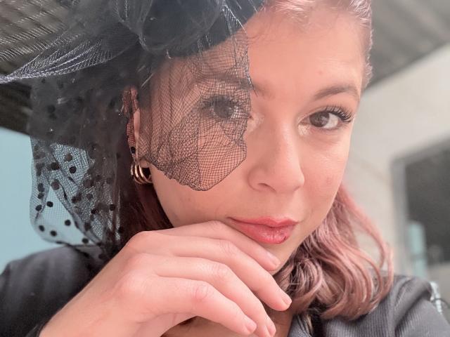 MoonXVelvet has pinkish-red hair, wearing a black lace headscarf, and is touching her chin with her hand, looking at the camera with a slight smile. Her lips are puckered.