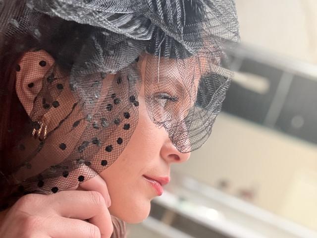 MoonXVelvet with a black polka-dot mesh veil, her face in profile, subtle smile, red lipstick, gold earring, and delicate hand resting near her cheek, looking out a window.