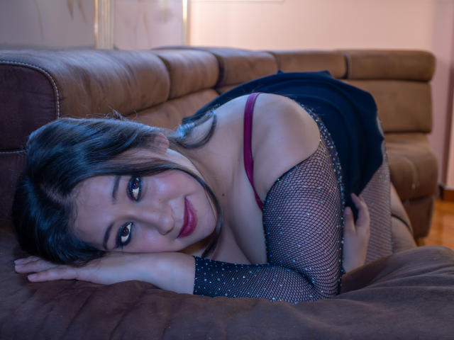 MelodyRoss, avec ses cheveux noirs et son soutien-gorge rouge, est assise sur un canapé marron, portant des manches en résille et une robe noire. Elle sourit à la caméra, montrant ses seins et ses fesses, dans une pièce peu éclairée..