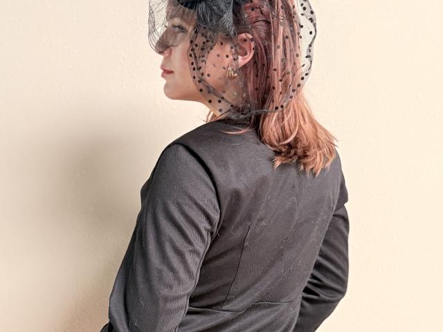 MoonXVelvet with pink-dyed hair, wearing a black, long-sleeved, shiny dress, and a black polka-dot, netted headpiece, looking to the side with hands on her back.