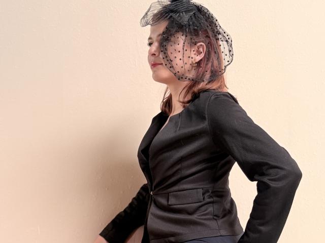 MoonXVelvet with a black polka-dot headscarf, wearing a black long-sleeve blazer, hands on hips, side profile, light skin, brown hair, subtle smile, wall in background.