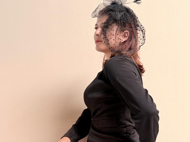 MoonXVelvet with small tits, wearing a black long-sleeve dress, and a black sheer polka-dot headpiece with a feather, standing sideways against a plain wall.