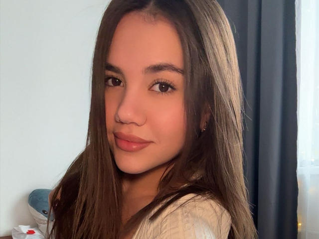 SantinaSchaumberg has long brown hair, a nose ring, and is smiling with pink lips. She's wearing a white top and has a cute, attractive face.