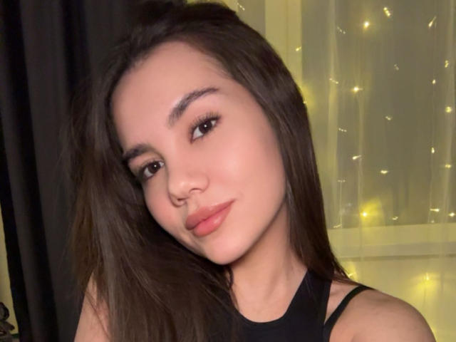 SantinaSchaumberg has long brown hair, a cute nose, and full pink lips. She's wearing a black top, looking sexy with a slight smile, and has subtle makeup.