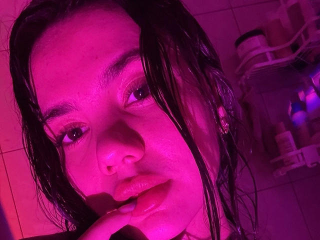 SantinaSchaumberg with wet black hair, pink lit face, finger near pink lips, looking into camera, purple light, tile background, soft expression, subtle makeup, small stud earring.