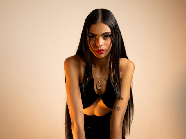 CrystalDavids, avec de longs cheveux noirs, du rouge à lèvres rouge, un haut noir découpé, des tatouages sur les bras, penchée en avant, un collier avec un pendentif ovale, sexy, peau brune, poitrine moyenne, lumière douce..