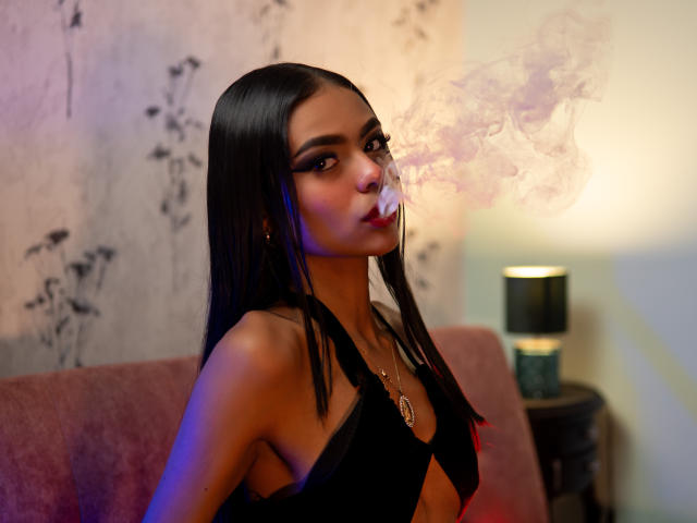 Crystal Davids, con lunghi capelli neri, che fuma uno spinello, soffiando fumo viola, indossa un top nero con spalline, mostrando il seno, e una collana d'oro, in una stanza poco illuminata..