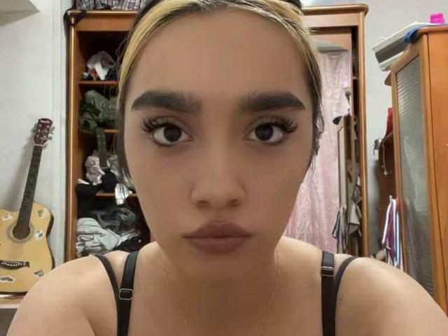 ShanaRoca, avec sa peau mate, ses sourcils foncés, ses longs cils et ses cheveux blonds, portant un haut noir à bretelles, regardant directement l'appareil photo avec des lèvres pincées..