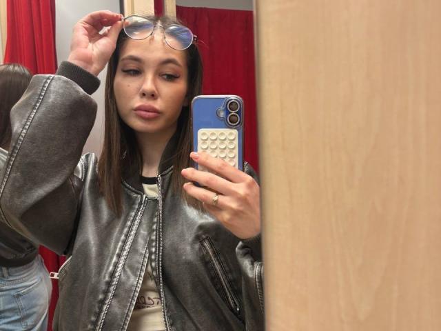 Karleen Tallas, com cabelo castanho, vestindo uma jaqueta de couro cinza, segurando um telefone azul com uma capa branca, abaixando os óculos, mostrando seus lábios curvados, em um selfie no espelho..