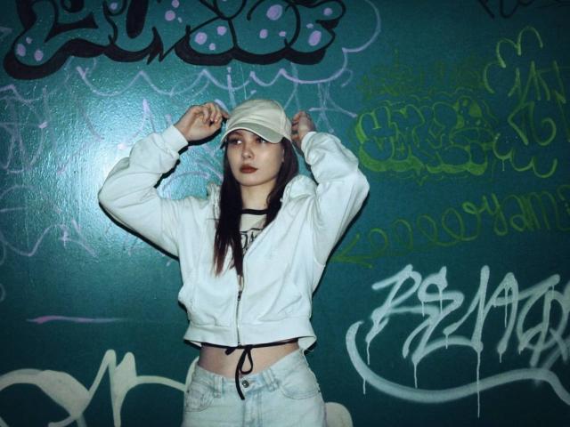Karleen Tallas, con lunghi capelli castani, indossa un berretto bianco, una giacca bianca, una collana nera e jeans chiari, è in piedi con le braccia alzate, e sullo sfondo si vede un muro verde ricoperto di graffiti..