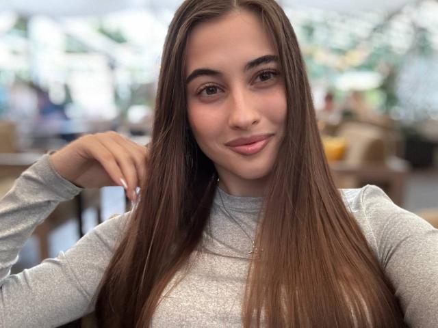 LateshaLuckenbaugh has long brown hair, a cute face, and small tits, wearing a gray long-sleeve shirt, smiling softly with her hand near her head in a selfie.