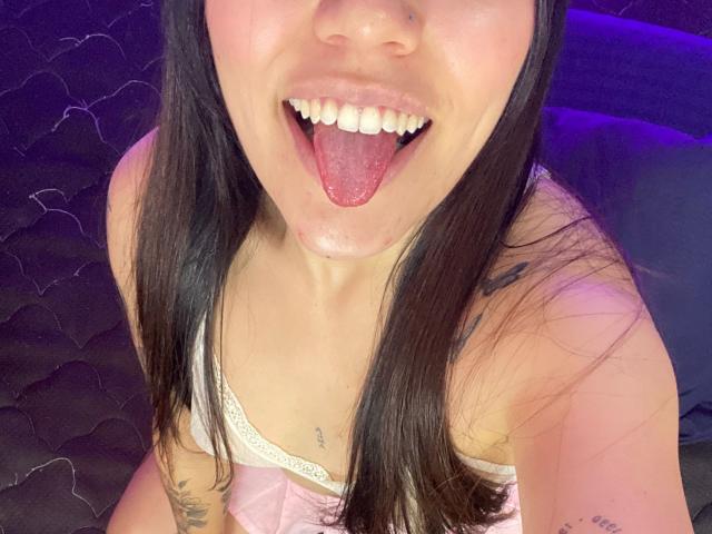 SophiaMarx with black hair, open mouth, tongue out, visible teeth, white tank top with lace, small tattoos, pink skin, sitting on dark quilted bed, purple light.