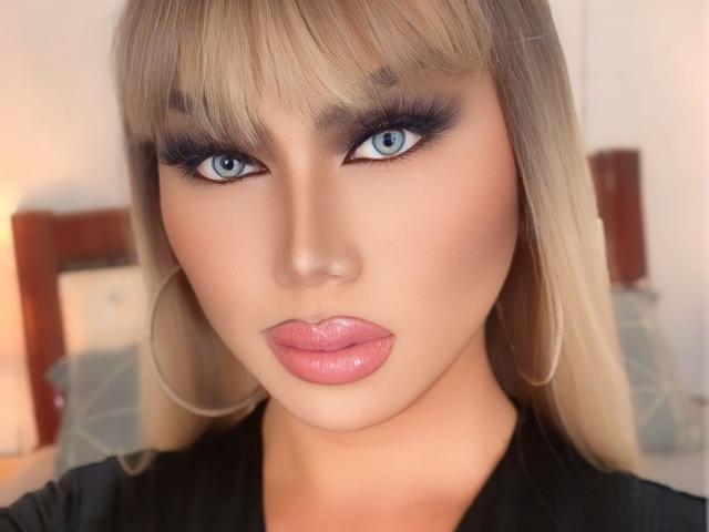 QueenDomVictoria has blonde hair, blue eyes, dark eyeliner, and pink glossy lips. She's looking directly at the camera with a serious expression, wearing a black shirt.
