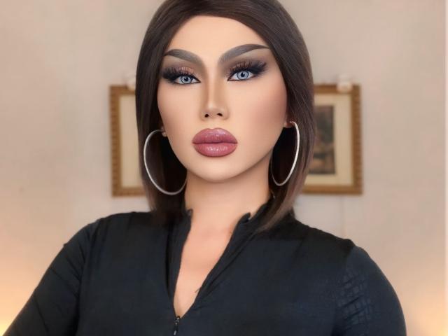 QueenDomVictoria with blue eyes, dark eyebrows, and pink lips, wearing a black zip-up shirt, large silver hoop earrings, and a sleek brown bob, looking directly at the camera.