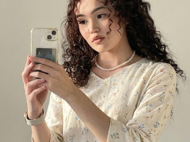 DebPlagens, com cabelos castanhos encaracolados, vestindo um vestido floral e um colar de pérolas brancas, tirando uma selfie com um celular dourado, usando um smartwatch, com uma expressão séria..