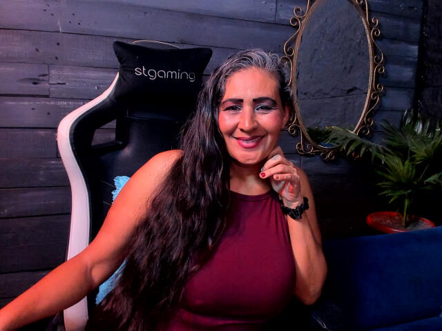 Victoria Kiley, con cabello castaño largo y ondulado, sonriendo, vestida con una blusa burdeos sin mangas, sentada en una silla de juego de estrellas negra y blanca, tocándose la barbilla. Sus senos son visibles..