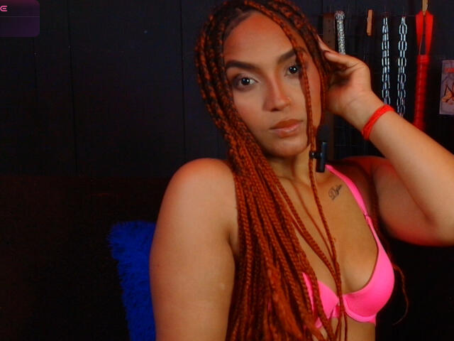 BrianaPratt with long red braids, pink bra, and red wristband, hand in hair, tattoo on right chest, looking at camera, dark background, blue pillow.