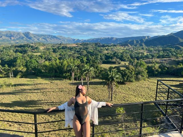 AlessiaFerry with big tits, wearing a black one-piece and white shirt, arms out, standing on a balcony, looking up, pink hairband, green hills and mountains in the background.