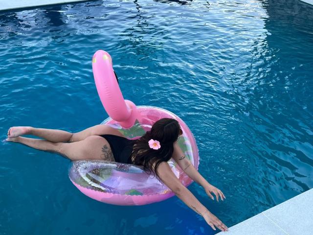 AlessiaFerry, with her black one-piece, is floating on a pink inflatable flamingo in a blue pool, arms out, legs spread, tattoo on her left ass cheek.