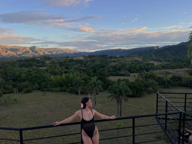 AlessiaFerry in a black one-piece swimsuit with high-cut sides, standing with arms on a black railing, looking at mountains under a blue, cloudy sky. Her pink hair tie.