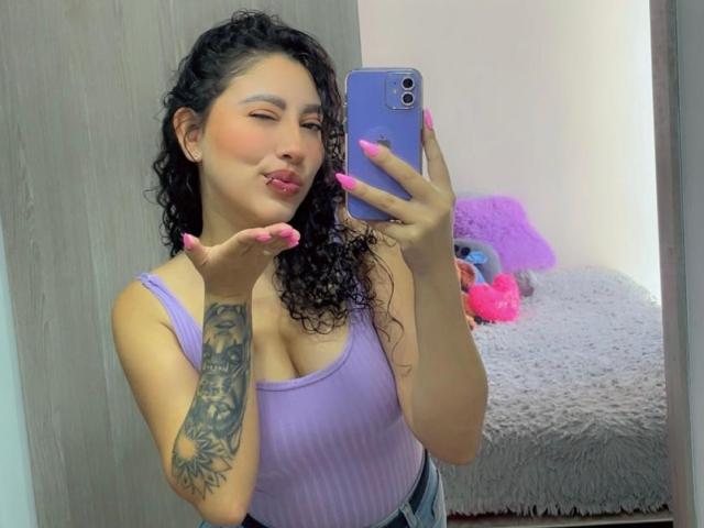 AmaraDeLux with curly black hair, pink nails, purple tank top, large tits, tattoo on left arm, pouty lips, holding purple phone, taking mirror selfie.