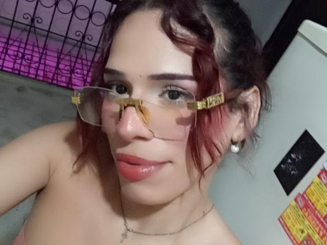 LucianaFlame with red curly hair, wearing clear glasses with gold "B-REPS" tags, pink lipstick, and pearl earrings, showing her bare shoulders and a subtle smile.