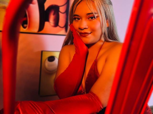 JadeyBrown, avec ses longs cheveux blonds, son eye-liner rouge et ses gants en satin rouge, touchant sa joue, portant un haut en dentelle rouge, souriant, montrant ses seins, dans une pièce éclairée en rouge..