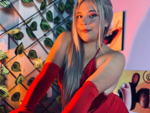 JadeyBrown, con lunghi capelli biondi, indossa un abito rosso a fascia e guanti rossi coordinati, è china in avanti, mostrando il seno, sorridendo con luci rosa e arancioni..