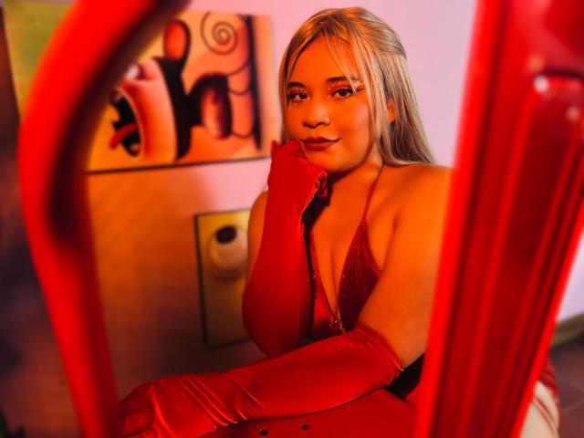 JadeyBrown, con i capelli biondi, indossa un abito rosso, lucido e con un profondo scollo, e guanti abbinati, posando con il mento appoggiato sulla mano, appare sensuale, in una stanza illuminata di rosso..