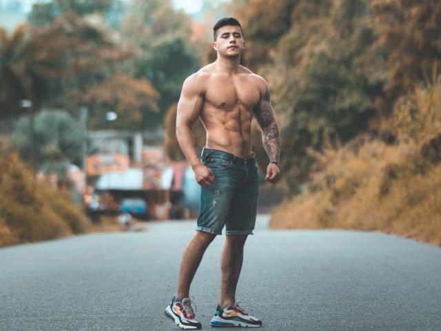 GarrettAlvin, musculoso, com tatuagens no braço direito, sem camisa, vestindo calças jeans verdes e tênis coloridos, em pé em uma estrada com folhagem de outono..