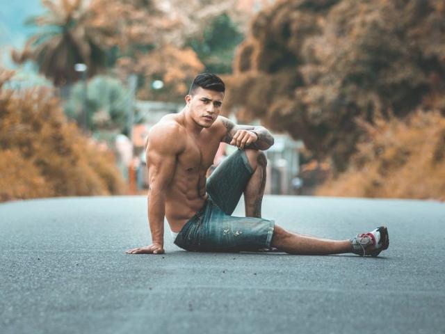 GarrettAlvin ha un corpo muscoloso e senza camicia, con addominali scolpiti, indossa jeans blu strappati e scarpe da ginnastica, ed è seduto su una strada con un'espressione seria..