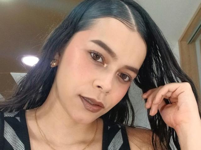 AlanaBlake has black hair, brown eyes, and dark lipstick. She's wearing a black and white striped top with a gold necklace. Her hand is in her hair.