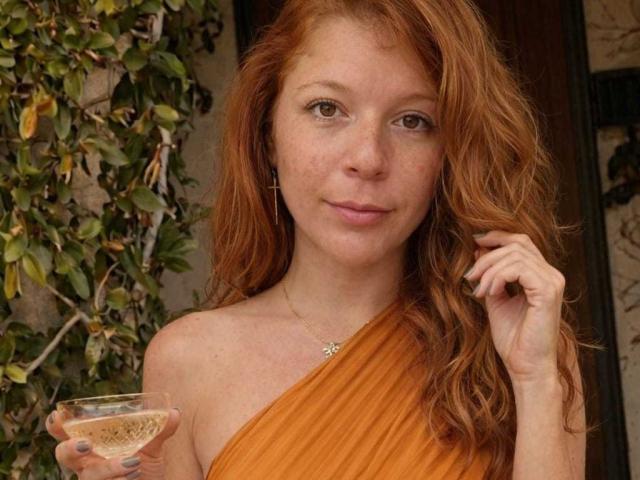 MoonXVelvet with red hair, wearing an orange one-shoulder dress, holding a glass of white wine, freckles on face, cross earrings, gentle smile, right hand touching hair.