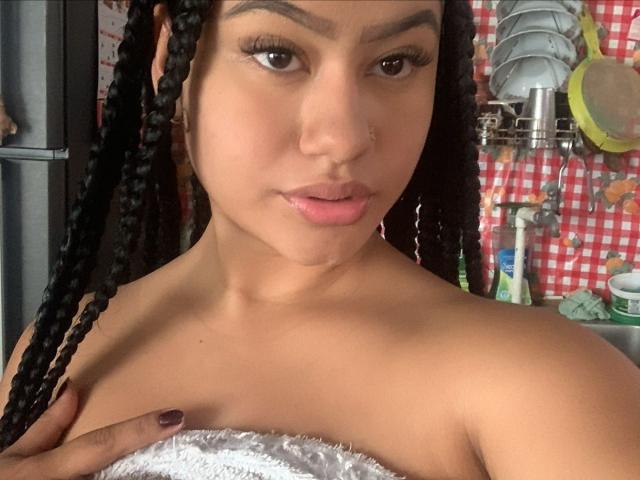 NoiraVale with braided black hair, nose piercing, and pink lips, holding a white towel over her bare tits, looking at the camera with dark eyes and purple nails.