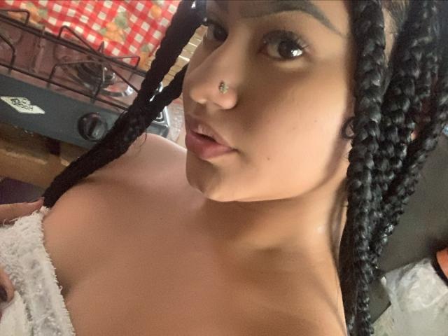 NoiraVale with dark braids, nose ring, and a slight pout, showing bare shoulders and a hint of her large, round tits. She has a light brown skin tone.