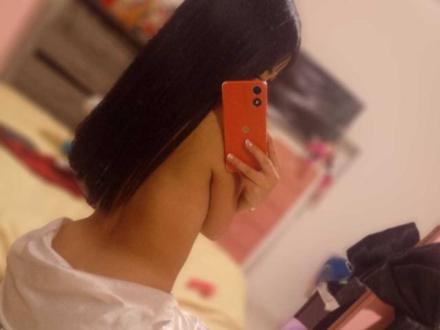 JulesDurand, topless with long black hair, holds an orange phone, covering her face, showing her bare back and white pants, in a blurry bedroom selfie.
