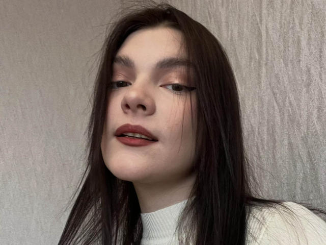 LatriceVer has pale skin, long straight brown hair, and is wearing red lipstick with black eyeliner. She's looking slightly to the side with a subtle smile.