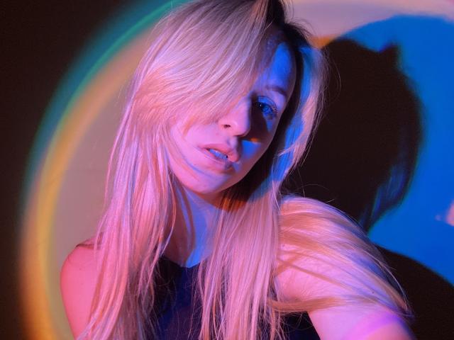 FlyFreedom met lang blond haar, roze en blauwe verlichting, schaduw op de muur, kijkt iets opzij, roze lippen, blauw oog, zachte uitdrukking, roze en blauw regenbooglicht effect..