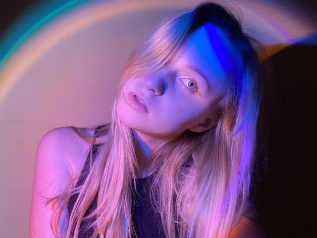 FlyFreedom ha i capelli biondi, gli occhi azzurri e una leggera inclinazione del capo. Indossa una canotta nera senza maniche, illuminata da luci viola e rosa con un delicato effetto arcobaleno..