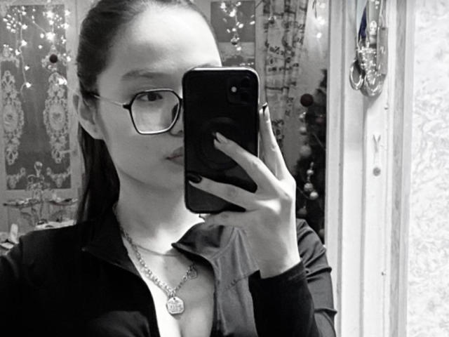 Black-and-white selfie of LessieMelendy with glasses, dark hair in a ponytail, wearing a zip-up shirt and necklace, holding a phone, showing her tits slightly.