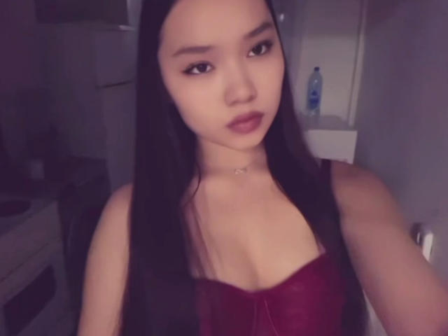 A selfie of LessieMelendy with long black hair, wearing a red corset that highlights her tits, and a subtle necklace, looking at the camera with dark lipstick.