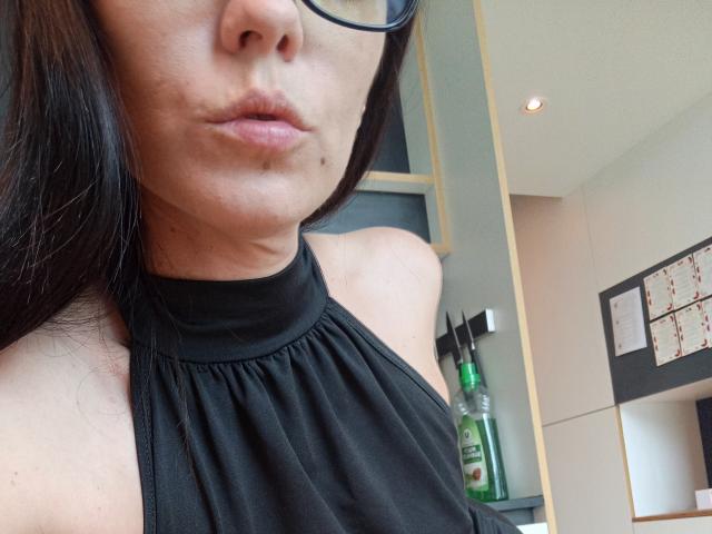 OrianaLaFrancaise with pouty lips, wearing black halter top, showing bare shoulders, black-framed glasses, and a small mole under her nose. Her dark hair falls over her face.