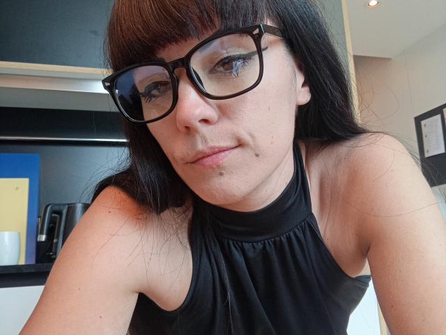 OrianaLaFrancaise with black-framed glasses, long brown hair, black sleeveless top, small moles on her face, and a subtle smile, showing her bare shoulders and chest.