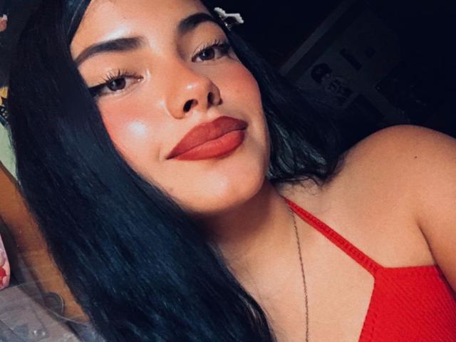 BrianaClarkX, con i suoi lunghi capelli neri, il rossetto rosso e la canotta rossa, mostra il suo viso luminoso e il suo adorabile sorriso, con il seno leggermente visibile, indossando una sottile collana..