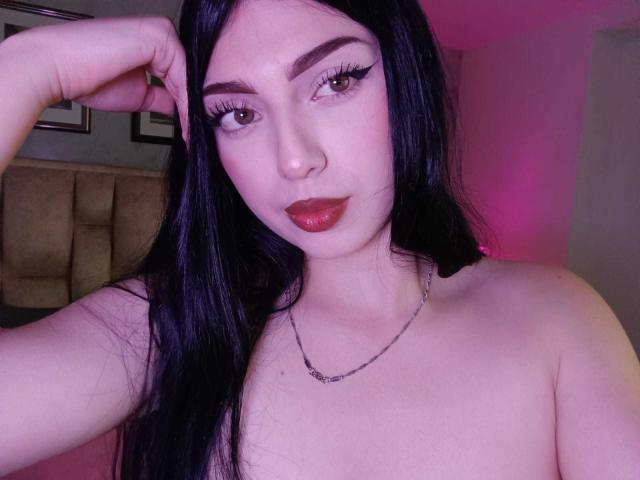 IsabellaaRossi with long black hair, pink eyeliner, and red lipstick, topless, showing her tits, hand in hair, pink and purple room lighting, necklace, serious expression, photo.