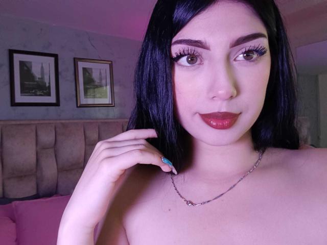 Isabella Rossi, con i capelli neri, il rossetto rosso e le lunghe ciglia, mostra il seno con una collana d'argento e un anello blu, in una camera da letto illuminata di rosa..