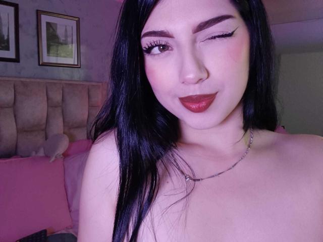 Isabella Rossi, com um olho piscando, sobrancelhas escuras, cabelo preto longo, batom vermelho e um colar de prata, mostrando os ombros nus, sorrindo e usando delineador preto. Seios visíveis na parte inferior da imagem..