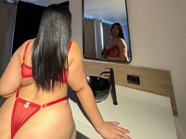 RashaelBig with long black hair, red lingerie, facing mirror, showing her back and red lace panties, her ass, and a tattoo on her right side. Reflection shows her tits.