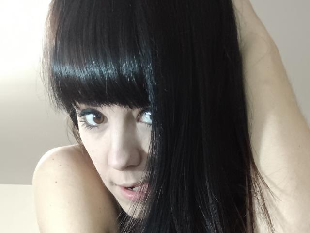 OrianaLaFrancaise tiene el cabello negro y liso con flequillo, ojos marrones, y está mirando hacia arriba con la boca ligeramente abierta, con un hombro desnudo visible. Su rostro está parcialmente en sombra..