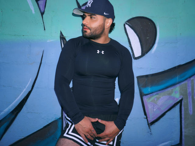 LyamVera draagt een strak zwart Under Armour shirt met lange mouwen, een zwarte pet met het NY-logo en zwarte korte broeken. Hij houdt zijn uitpuilende zwarte broek vast en staat voor een muur met graffiti..